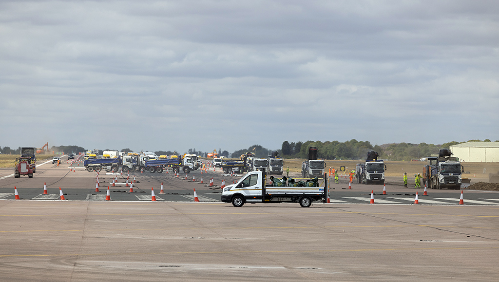A white flatbed van stands in the foreground of runway resurfacing project. Numerous construction trucks, dumpers, and road planning machines are lined up across the wide asphalt airfield, long into the distance, under an overcast sky, sectioned off by orange and white traffic cones.